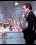 Man in a suit observing small alien creatures in a kitchen with a microwave, coffee pots, and mugs.