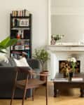 Elegant living room with a gray sofa, wooden chair, bookshelf, ornate mirror, and white fireplace with decor.