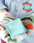 Hands in colorful knit mittens holding a small blue fabric sachet with a snowflake design.