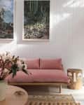 Pink cushioned sofa with wooden frame, floral arrangement on table, cactus wall art, and rustic side table in a bright room.