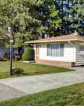 Single-story house with beige siding, large front window, attached garage, and manicured lawn surrounded by trees.