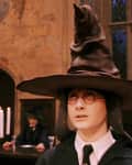 Young person wearing a large pointed hat in a medieval-style hall with candles and seated figures in the background.