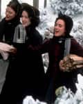 Four women in winter clothing carrying food and firewood through a snowy landscape.