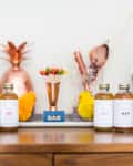 Bottles of botanical drinks on a wooden bar with a copper pineapple, cocktail tools, and wine rack.