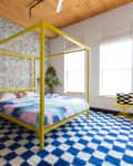 A bed with a yellow canopy frame, blue checkered rug, and pattered colorful wallpaper.