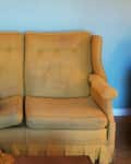 Yellow vintage sofa with button-tufted backrest against a blue wall, framed art above.