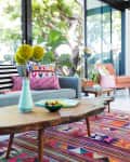 Living room with a gray sofa, colorful pillows, wooden coffee table, and vibrant geometric rug near large windows.