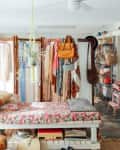 Bohemian bedroom with floral bedspread, hanging plants, vintage fans, and a room divider displaying clothes and bags.