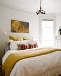 Cozy bedroom with a white bed, yellow accents, colorful pillows, a dresser, and abstract wall art.
