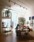 Loft-style art studio with large windows, plants, and colorful paintings on the walls. Two people working at a central table.