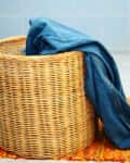 Wicker laundry basket with blue jeans draped over, on an orange rug in a tiled room.