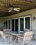 Covered patio with ceiling fan, glass table, and cushioned chairs, next to sliding glass doors.