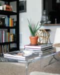 Modern living room with clear coffee table, books, potted plant, white chair with fur throw, and black bookshelf.