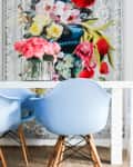 White dining table with blue chairs, pink flowers in a vase, and a colorful floral painting in the background.