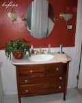 Bathroom with red walls, wooden vanity, oval mirror, gold fixtures, and a potted plant.