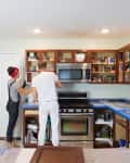Couple renovating a kitchen, organizing open cabinets above a stainless steel stove and microwave.