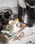 Coffee station with Keurig machine, white mug on tray, coffee pods, and jar of beans on a granite countertop.