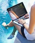 Person sitting by a pool using a laptop, feet in the water, wearing a sleeveless white top and dark shorts.