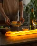 Person setting table with glowing baguette lamp, wine, salad, and rolls.