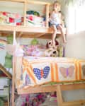 Child sitting on a wooden loft bed with colorful butterfly quilt, surrounded by stuffed animals and bright decor.