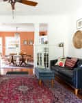 Living room with dark sofa, colorful cushions, red patterned rug, and view into dining room with orange walls and wooden table.