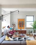 Modern living room with a gray sectional, geometric rug, wall art, and a person reading on a chair near a large plant.