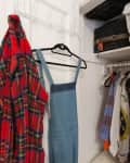 Small closet with hanging plaid shirt, denim overalls, and various clothes. Luggage stored on upper shelf.