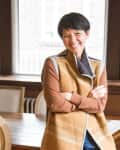 Person smiling in a dining room with wooden table, chairs, and a bowl of apples.