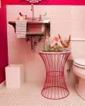 Bright pink bathroom with white tiles, pedestal sink, toilet, and a wire table holding decor and a striped towel.