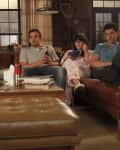 Four people sitting on brown leather sofas in a cozy living room, watching TV, with books and a lamp nearby.