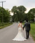 Bride in white gown and groom in gray suit walking hand in hand on a country road lined with trees.