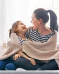 Mother and daughter in matching striped shirts laughing under a beige blanket on a bed.