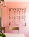 Wall-mounted calendar on pink wall above wooden chairs and white table.