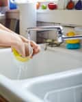 Person washing a yellow-striped cup in a kitchen sink with running water, surrounded by colorful kitchen items.
