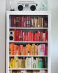 Bookshelf with color-coordinated books, a stereo, and a fan on top, next to a green plant against a white brick wall.