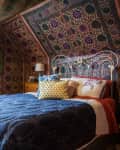 Eclectic bedroom with patterned wallpaper, blue quilt, ornate white headboard, and colorful pillows.