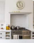 Modern kitchen with white cabinets, stainless steel stove, wall clock, and a ladder shelf holding a jug and candles.