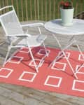 White patio chair and table on a deck with a red rug featuring white square patterns, topped with a potted plant.