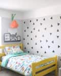Kids' room with yellow bed, triangle-patterned wall, round mirror, and colorful bedding. Decor includes animal toys and art.