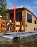 Wooden houseboat with a deck on a lake, surrounded by trees and a sandy shore.