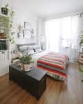 Cozy bedroom with a bed, striped blanket, plants, guitar, and natural light from sheer curtains.