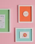 Three framed artworks on a pink wall: green with text, orange with circular design, and blue with spaghetti illustration.