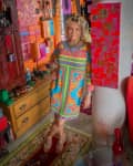 Woman in a colorful dress standing in a vibrant room with eclectic art and decor.