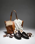 Brown leather tote with fur-trimmed gloves, cozy scarf, and black fur-lined slippers surrounded by pinecones.