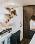 Two people cooking in a cozy kitchen, one stirring a pot on the stove, the other chopping vegetables at the sink.