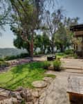 Mid-century modern patio with wooden bench, stone path, lush greenery, and view of distant hills.
