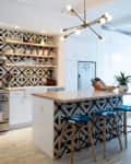 Modern kitchen with geometric backsplash, open shelves, island with stools, and pendant light. Living area visible in background.