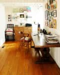 Home office with wooden desk, chairs, wall art, and shelves with books and decor in a bright, narrow room.