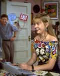 Teen girl in a colorful top using a computer, with a boy leaning on a door marked "Exit" in the background.