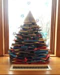 Stacked books forming a Christmas tree with red lights and a broom on top, set on a wooden windowsill.
